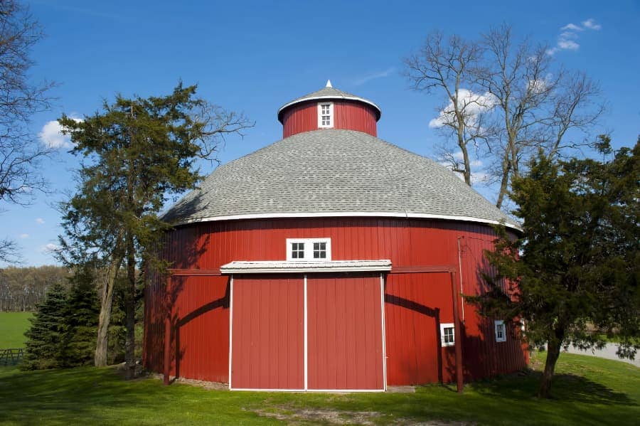 Conical shaped, red walled barn sitting together with green trees and blue sky